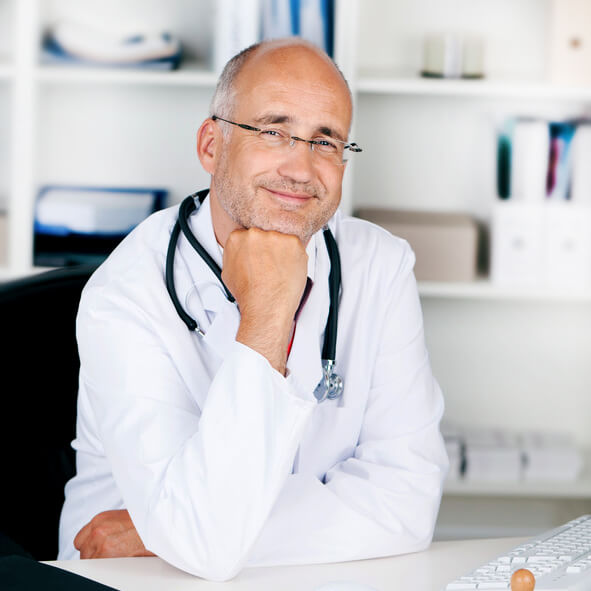 A smiling doctor with a stethoscope around his neck sitting at a desk in a medical office.