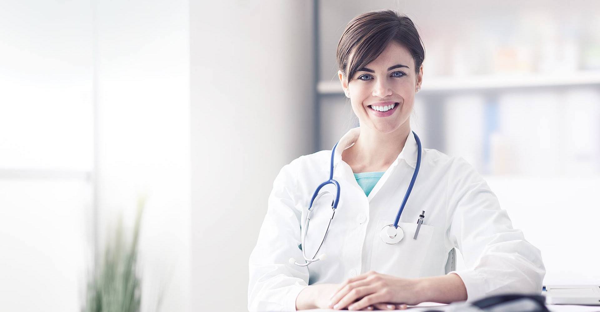 Female healthcare professional with a stethoscope smiling at the camera in a clinical setting.